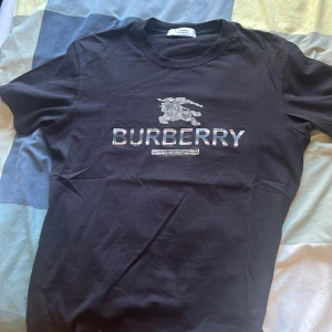 Svart Burberry t-shirt med logga - Oanvänd Snygg svart t-shirt från Burberry med klassisk logga och emblem tryckt på bröstet. T-shirten har rund hals och korta ärmar, perfekt för dig som gillar stilrena märkesplagg. Materialet känns mjukt och skönt mot huden.