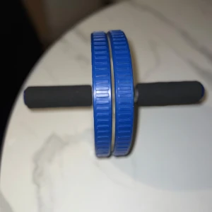 Träningsutrustning  - Bilderna visar en träningsutrustning, en maghjul/ab roller med blå hjul och svarta handtag. Perfekt för dig som vill träna core och bygga styrka hemma! Enkel att använda och passar både nybörjare och erfarna. Gör din träning roligare och utmana dig själv med denna smidiga träningspryl.