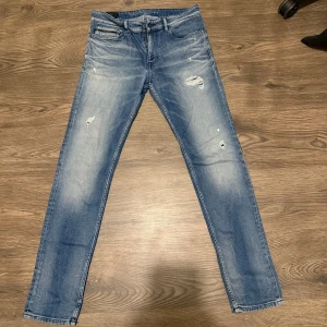 Calvin Klein skinny jeans blå W31 L32 - Snygga blå skinny jeans från Calvin Klein i premium italienskt tyg. Modellen har slitningar och hål på framsidan för en cool, trendig look. Klassisk femficksdesign och smal passform som sitter tajt längs benen. Perfekt för dig som gillar streetstyle och vill sticka ut.