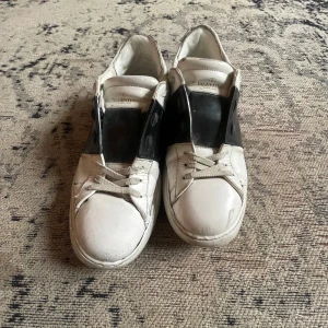 Valentino Garavani Open sneakers vit/svart - Säljer ett par Valentino Garavani Open sneakers i vit skinn med bred svart läderpanel över mitten. Klassisk rund tå, platt sula och snörning framtill. Snygga nitar på hälen ger extra edge. Perfekt för dig som gillar exklusiva street vibes.