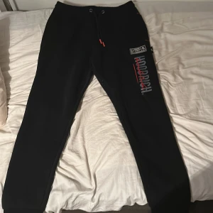 Svarta mjukisbyxor från Hoodrich - Svarta mjukisbyxor från Hoodrich med tryck och broderade detaljer i vitt och rött. Byxorna har dragsko i midjan och ribbade muddar vid bensluten. Snygg streetstil med logga på benet och coola detaljer som sticker ut.