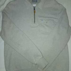 Beige tröja med half zip från Morris - Snygg beige tröja från Morris med krage och half zip-dragkedja med läderdetalj. Bröstficka med knapp och ljusblå broderad logga. Mjuk bomullskvalitet och långa ärmar, perfekt för chill höst- och vårväder. I väldigt gott skick.