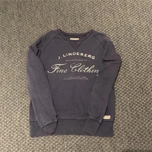 Mörkblå sweatshirt från J.Lindeberg - Snygg mörkblå sweatshirt från J.Lindeberg med tryck på bröstet och en liten tygpatch vid nederkanten. Tröjan har rund hals och långa ärmar, perfekt för chill dagar. Mjuk bomullskänsla och klassisk passform. Passar typ 170 till 180