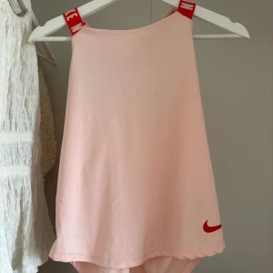 Ljusrosa Nike baddräkt med röda band - Supersnygg ljusrosa baddräkt från Nike med breda röda axelband där det står NIKE i vitt. Baddräkten har en hög halsringning och korsade band i ryggen för extra cool look. Materialet är mjukt och stretchigt syntet, perfekt för strand eller pool.