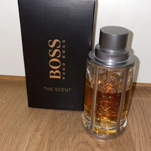 Hugo Boss The Scent 100ml EdT - Hugo Boss The Scent är en elegant herrparfym i en stilren glasflaska med silverdetaljer och bärnstensfärgad vätska. Doften har inslag av ingefära, exotisk maninka och läder. Flaskan rymmer 100 ml och kommer i en svart kartong med kopparfärgade detaljer. Som man ser på flaskan så är den inte full men mer en halva e kvar. Ny pris är runt 650kr. 