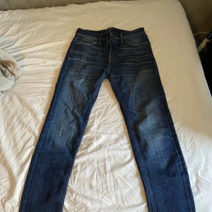 Blå tapered jeans från Jack & Jones - Snygga blå jeans från Jack & Jones med tapered fit och klassisk femficksdesign. Jeansen har en mörk tvätt med lätt slitning och kontrastsömmar. Modellen heter MIKE och är tillverkad i jeansmaterial med dragkedja och knappstängning. Perfekta för en chill och stilren look. Hör av er för mer info!