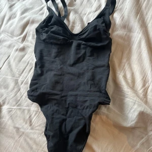 Svart body med justerbara band - Snygg svart body från Skims med justerbara axelband och lätt v-ringad hals. Modellen är enkel och stilren, perfekt för dig som gillar en klassisk look på stranden eller vid poolen. Materialet är mjukt och följsamt för en bekväm passform.