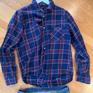 Rutig blå och vinröd skjorta från 157 - Säljer en rutig skjorta från 157 i storlek M. Skjortan har en schysst kombination av blått och vinrött med vita linjer, klassisk krage och bröstficka. Långärmad och tillverkad i mjuk bomull, perfekt för chill dagar. Knappar framtill och på ärmsluten.