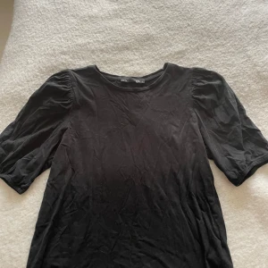 Svart topp med puffärm från H&M - En svart topp med rund halsringning och trendiga puffärmar som ger en snygg siluett. Toppens material är mjuk bomull och den har en enkel, stilren look som passar till många outfits. Perfekt för dig som gillar minimalistisk stil med en twist.