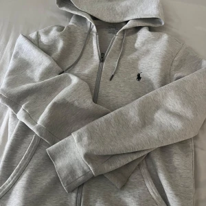 Grå hoodie från Polo Ralph Lauren - Snygg ljusgrå hoodie från Polo Ralph Lauren med dragkedja och huva. Tröjan har långa ärmar, snörning vid huvan och klassisk Polo-logga broderad på bröstet. Tillverkad i mjuk bomullsmix som är perfekt för chill eller häng. Passar dig som gillar stilren och enkel design. Säljs pågrund av att den är förstor och går ej att lämna tbx! (Självklart äkta)! Tveka inte på att höra av er vid minsta fråga! Fraktar samma dag!