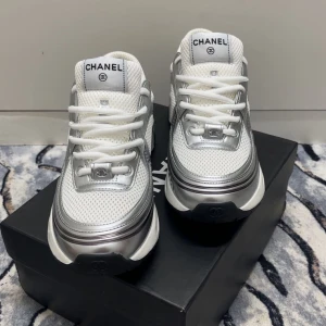 Chanel sneakers i vitt och silver - Snygga Chanel sneakers med chunky plattformssula i vitt, silver och svart. Ovandelen är i mesh och syntet med silvriga detaljer och tydligt Chanel-logo på sidan och tungan. Skorna har snörning och en rund tå, perfekt för dig som gillar lyxiga street vibes. Priset kan diskuteras 