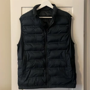 Marinblå dunväst från Zara - Snygg marinblå dunväst från Zara med quiltad design och dragkedja framtill. Västen har hög krage och två fickor på sidorna. Perfekt att slänga över en skjorta eller hoodie för en chill och stilren look.