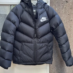 Svart pufferjacka från Nike stl 147-158 - Svart pufferjacka från Nike med huva och feta, quiltade sektioner. Jackan har dragkedja framtill, två sidofickor och stor Nike-logga på bröstet. Perfekt för kalla dagar och har en clean, sportig vibe. Passar dig som gillar streetwear och vill hålla dig varm.