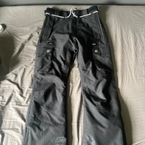 Svarta täckbyxor från Dope - Svarta täckbyxor från Dope med coola detaljer som snörning i midjan och stora fickor med tryckknappar. Byxorna har bred passform och är gjorda i slitstark polyester, perfekta för vintern och snöaktiviteter. Snyggt Dope-märke på benet och insidan.