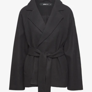  Gina Tricot kappa med bälte - Svart kappa från Gina Tricot i omlottmodell med bälte i midjan. Jackan har klassisk krage, långa ärmar och två fickor framtill. Materialet är mjukt och känns lyxigt, perfekt för dig som gillar stilrena och tidlösa plagg. Snygg att matcha med boots eller sneakers.