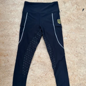 Svarta ridleggings från Maya Delorez - Svarta ridleggings från Maya Delorez med hög midja och vita sömmar. Snygga silikonmönster på insidan av benen för extra grepp i sadeln. Tillverkade i stretchigt syntetmaterial som andas och sitter tajt. Perfekta för dig som vill ha både stil och funktion i stallet.