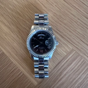 Silverfärgad Lotus herrklocka - Stilren klocka från Lotus i silverfärgat rostfritt stål med svart urtavla. Klockan har dag- och datumvisning, tydliga index och romerska siffror. Länkarmbandet är robust och har Lotus-loggan ingraverad på spännet. Perfekt accessoar för dig som gillar klassisk design.