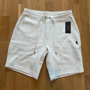 Grå shorts från Polo Ralph Lauren - Snygga ljusgrå shorts från Polo Ralph Lauren med klassisk logga broderad i mörkblått. Mjukt bomullsmaterial, elastisk midja med snörning och praktiska fickor fram samt en bakficka. Perfekta för chill dagar eller när du vill ha en clean look.