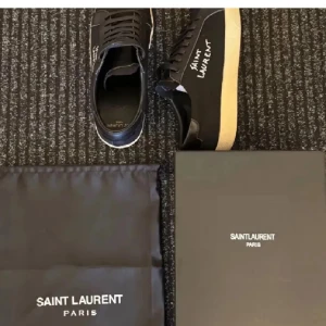 Svarta Saint Laurent sneakers - Svarta sneakers från Saint Laurent med vit logotyp på sidan och beige platt sula. Skorna har snörning och är tillverkade i en kombination av skinn och textil, vilket ger en snygg kontrast. Perfekt för dig som gillar stilrena och exklusiva sneakers.
