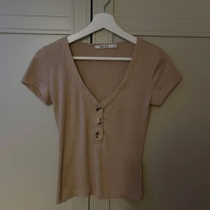 Beige ribbad topp med knappar NA-KD - Säljer en beige ribbad topp från NA-KD i storlek XS. Toppen har korta ärmar, v-ringning och tre snygga knappar framtill. Perfekt för dig som gillar en enkel men trendig stil. Materialet är mjukt och stretchigt för en skön passform.