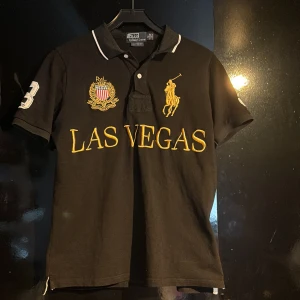 Svart Polo Ralph Lauren Las Vegas tröja - Svart t-shirt från Polo Ralph Lauren med guldig broderad text 'LAS VEGAS' och logga på bröstet. Stor vit siffra 3 på ryggen och ärmen. Klassisk pikékrage med vit kant, custom fit. Materialet är mjuk bomull, perfekt för en chill och sportig look.