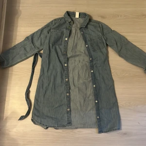 Blå jeansskjorta med bälte från &Denim - Snygg lång jeansskjorta från &Denim i ljusblå denim med två bröstfickor, knappar framtill och tillhörande bälte i midjan. Skjortan har klassisk krage och långa ärmar, perfekt för en chill och avslappnad look.