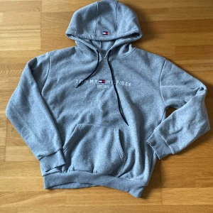 Grå hoodie Tommy Hilfiger L - Säljer en grå hoodie från Tommy Hilfiger i storlek L. Tröjan har broderad logga och text på bröstet, klassisk känguruficka och svarta snören i huvan. Mjuk och skön bomullsmix, perfekt för chill dagar. Ribbad mudd vid ärmslut och nederkant.