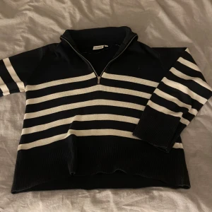 Randig stickad tröja Kappahl - Snygg stickad tröja från Kappahl i mörkblått och vitt med breda horisontella ränder. Tröjan har en hög krage och en half zip-dragkedja framtill. Långärmad med breda muddar och skön passform. Perfekt för dig som gillar klassisk stil med en twist.