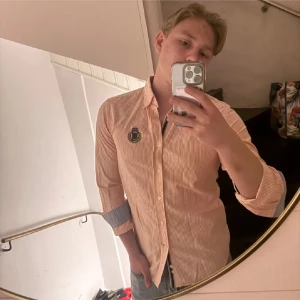 Randig skjorta Morris Stockholm M - Snygg randig skjorta från Morris Stockholm i storlek M. Skjortan har smala ränder i beige och vit, klassisk krage, långärmad design och knappar framtill. Broderad patch på bröstet och kontrastfärgade manschetter. Tillverkad i mjuk bomull med Twofold Yarn. 249kr st eller 399kr för båda