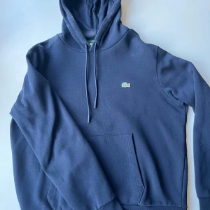 Marinblå hoodie från Lacoste - Säljer en stilren marinblå hoodie från Lacoste med klassisk krokodillogga på bröstet. Tröjan har huva med snörning, stor magficka och långa ärmar. Perfekt för chill dagar och enkel att matcha med jeans eller joggers.