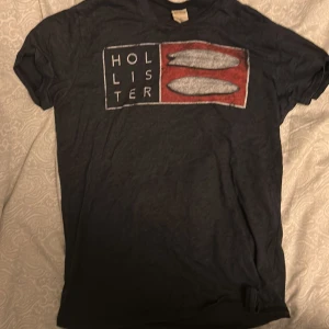 Svart Hollister t-shirt med tryck - Svart t-shirt från Hollister i storlek S med ett stort grafiskt tryck på bröstet i rött, vitt och grått. Trycket har surfbrädor och texten 'Hollister'. Klassisk rund hals och korta ärmar. Mjuk bomullskvalitet som är skön mot huden.