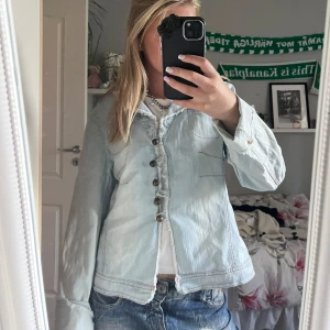 Ljusblå jeansjacka med knappar - Säljer en ljusblå jeansjacka med coola metallknappar framtill och råa kanter vid kragen. Jackan har en avslappnad passform och långa ärmar, perfekt att slänga över en t-shirt. Klassisk denimlook som funkar till jeans eller kjol. vintage