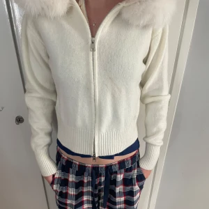 Alessa fur jacket - Snygg vit cardigan jacka med dragkedja framtill och en stor fluffig huva med pälsdetalj. Jackan är stickad och har ribbade muddar vid ärmslut och nederkant. Perfekt för dig som gillar mysiga och trendiga plagg med lite extra attityd.