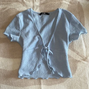 Ljusblå omlottopp från Bik Bok - Supersöt ljusblå omlottopp från Bik Bok med korta ärmar och knyt framtill. Toppen har en v-ringad design och är gjord i ett mjukt, ribbat material som ger en chill och feminin vibe. Perfekt att styla med jeans eller kjol för en fräsch look.
