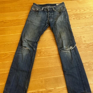 Blå slitna jeans från Diesel - Säljer ett par blå jeans från Diesel med coola slitningar och hål på knäna. Jeansen har klassisk femficksdesign, rak passform och snygga detaljer som fransade benslut och slitna bakfickor. Perfekt för dig som gillar en avslappnad och edgy stil. W31 L34