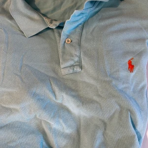 Ljusblå pikétröja från Polo Ralph Lauren - Snygg ljusblå pikétröja från Polo Ralph Lauren i storlek L. Klassisk krage, två knappar och den ikoniska orange broderade loggan på bröstet. Tröjan är gjord i mjuk bomull och har en stilren, normal passform som funkar perfekt till jeans eller shorts.