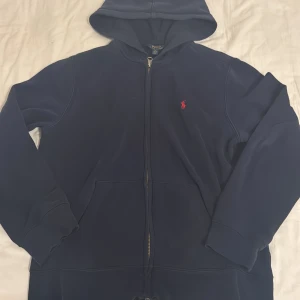 Marinblå hoodie från Polo Ralph Lauren - Snygg marinblå hoodie från Polo Ralph Lauren med dragkedja och huva. Klassisk röd logga broderad på bröstet. Tröjan har långa ärmar, två fickor fram och är gjord i mjuk bomullsmix som är skön att ha på sig. Perfekt för chill dagar eller när du vill ha en clean look.