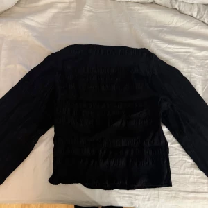Svart långärmad blus med struktur - Säljer en svart blus med lång ärm och snyggt strukturerat tyg. Modellen är rak och har en lätt rynkad effekt över hela plagget, vilket ger en cool och modern vibe. Perfekt att styla med jeans eller kjol för en trendig look.