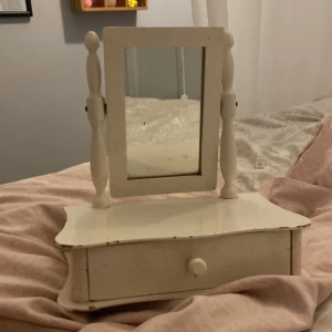 Vit sminkspegel med låda - Söt vit sminkspegel i trä med klassisk design och svängbar spegel. Spegeln har två dekorativa sidostolpar och en praktisk låda under för förvaring av smink eller accessoarer. Perfekt storlek för sminkbordet och ger en vintagekänsla till rummet.