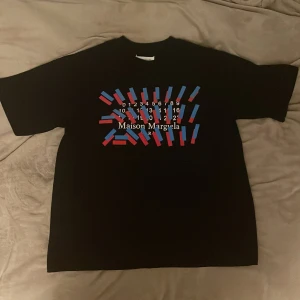 Maison Margiela t-shirt - Svart t-shirt från Maison Margiela med en fet grafiskt print i rött och blått på bröstet och med deras vita logga. Klassisk rund hals och korta ärm. Passformen är loose och t-shirten har en clean look med diskreta vita stygn bak i nacken. Fick den av min brorsa men jag tycker den är för baggy för min del
