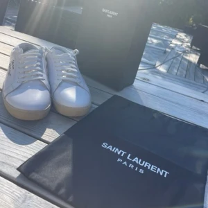 Saint Laurent vita sneakers i skinn - Snygga vita sneakers från Saint Laurent med handskriven logga på sidan. Skorna har rund tå, platt sula och klassisk snörning. Tillverkade i lyxigt skinn med beige gummisula. Perfekta för dig som gillar stilrena och exklusiva detaljer. Inget kvitto me  skorna är äkta. Dustbag och låda medkommer med skorna.