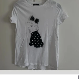 Vit t-shirt med pärlor och rosett - Vit t-shirt från Valley med korta ärmar och rund hals. Framsidan har ett tryck av en figur med svart rosett och kjol dekorerad med pärlor och prickigt mönster. Materialet känns mjukt och är troligen bomull. Perfekt för dig som gillar unika detaljer.