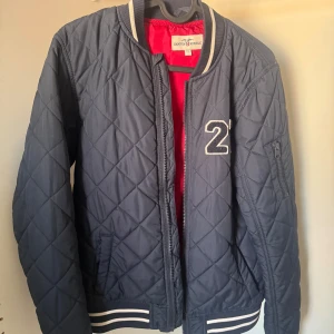 Mörkblå quiltad bomberjacka Hampton Republic stl. 170 - Quiltad mörkblå bomberjacka från Hampton Republic med coolt 27-tryck på bröstet och röd insida. Jackan har ribbade muddar med vita ränder, dragkedja framtill och fickor på sidorna. Perfekt för dig som gillar sportig och stilren look.