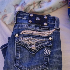 Miss Me jeans med broderi och strass - Säljer ett par blå Miss Me jeans med snygga broderade detaljer och strass på bakfickorna. Jeansen har kontrastsömmar, dekorativa knappar och ett coolt märke bak i midjan. Modellen har normal passform och klassisk jeanslook med extra bling.