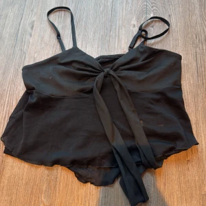 Svart linnetopp med knyt från Romwe - Svart linnetopp från Romwe med tunna justerbara axelband och snygg knytning framtill. Toppen har en lätt vågig nederkant och är tillverkad i ett tunt, luftigt syntetmaterial som känns skönt mot huden. Perfekt för att skapa en chill och trendig look.