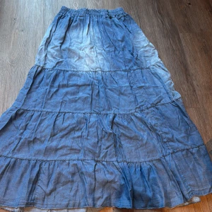 Blå långkjol med volanger - Säljer en blå långkjol med flera volanger och resår i midjan. Kjolen har en luftig och flowy känsla, perfekt för dig som gillar avslappnad stil. Materialet är mjukt och lätt, och färgen är en snygg denimblå ton.