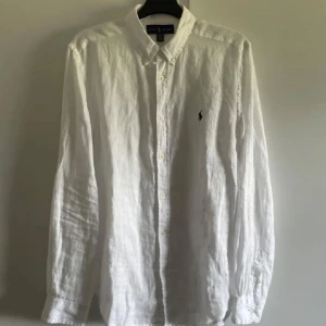 Vit linneskjorta från Ralph Lauren - Säljer en stilren vit linneskjorta från Ralph Lauren med klassisk krage och knappar framtill. Skjortan har långa ärmar och en broderad logotyp på bröstet. Perfekt för en avslappnad men ändå elegant look.