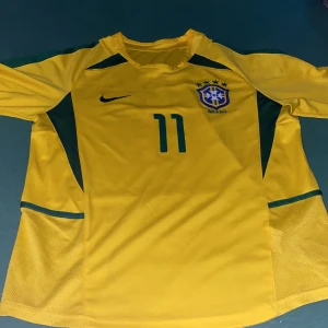 Ronaldinho 2002 vm. - Säljer en gul Brasilien fotbollströja från Nike med nummer 11 och texten 'Ronaldinho' på ryggen. Tröjan är kort ärmat och det brasilianska fotbollsförbundets emblem på bröstet. Perfekt för fotbollsfans! Tröjan är riktigt fin för sommaren och har bra kvalité.