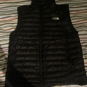 Svart dunväst från The North Face - Säljer en svart dunväst från The North Face. Västen har en dragkedja framtill och är quiltad för extra värme. Perfekt för kyliga dagar och har en stilren design med logotyp på bröstet.