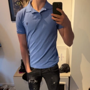 Blå pikétröja från Ralph Lauren - Säljer en snygg blå pikétröja från Ralph Lauren i storlek S. Tröjan har en klassisk krage och korta ärmar, med den ikoniska loggan broderad på bröstet. Perfekt för en stilren och avslappnad look.